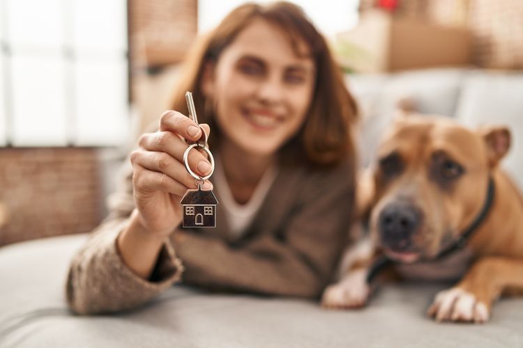 Junge kaukasische Frau hält den Schlüssel eines neuen Hauses und liegt mit ihrem Hund auf dem Sofa. Junge kaukasische Frau hält den Schlüssel eines neuen Hauses und liegt mit ihrem Hund auf dem Sofa.